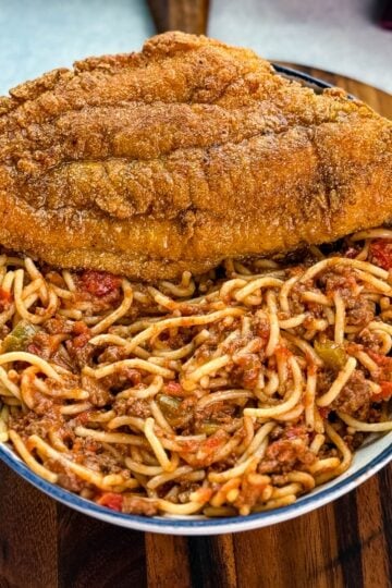 Fried catfish served with spaghetti on a white plate, garnished with shredded cheese, all placed on a wooden board.