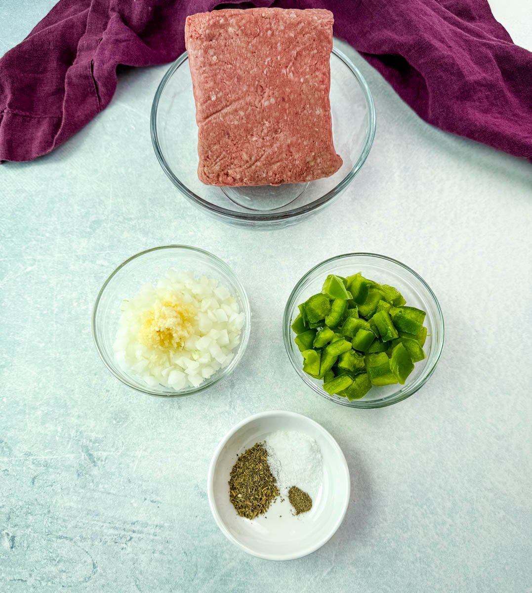 Overhead view of prepared spaghetti ingredients including raw ground meat in a bowl, chopped onions with minced garlic, diced green bell peppers, and a bowl of dry seasonings, all arranged on a light blue surface.
