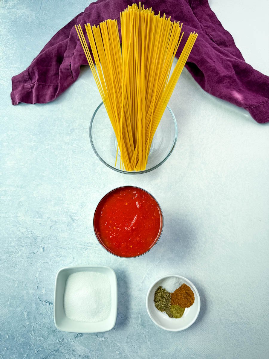Flat lay of uncooked spaghetti noodles in a glass bowl, canned crushed tomatoes, granulated sugar, and a bowl of seasonings (oregano, Cajun seasoning, and more) arranged neatly for making Southern-style spaghetti.