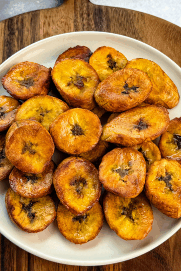 Plate of golden, crispy fried plantains stacked on a cream-colored plate set over a wooden board.