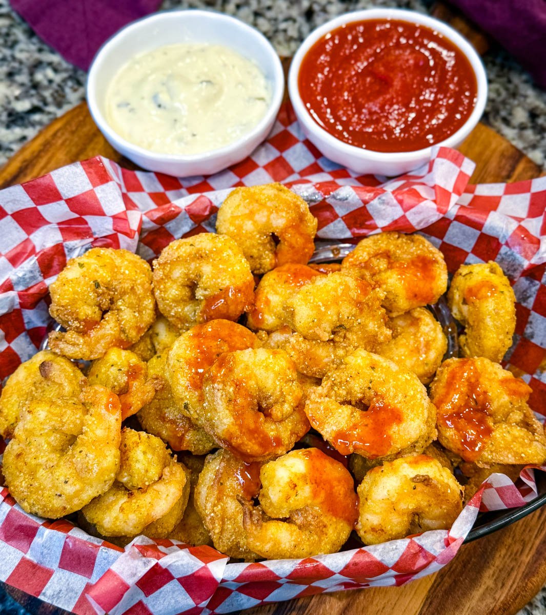 Close-up of crispy breaded fried shrimp in a basket, some topped with a drizzle of hot sauce, with dipping sauces blurred in the background.