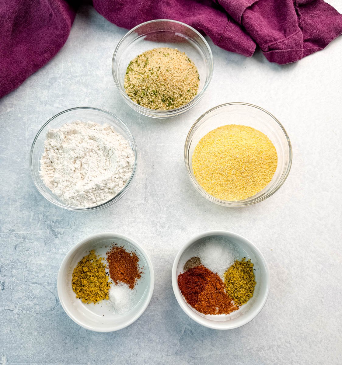 Overhead view of six small glass bowls showing individual ingredients for breading: flour, cornmeal, breadcrumbs, and seasonings, arranged on a light background with a purple towel.