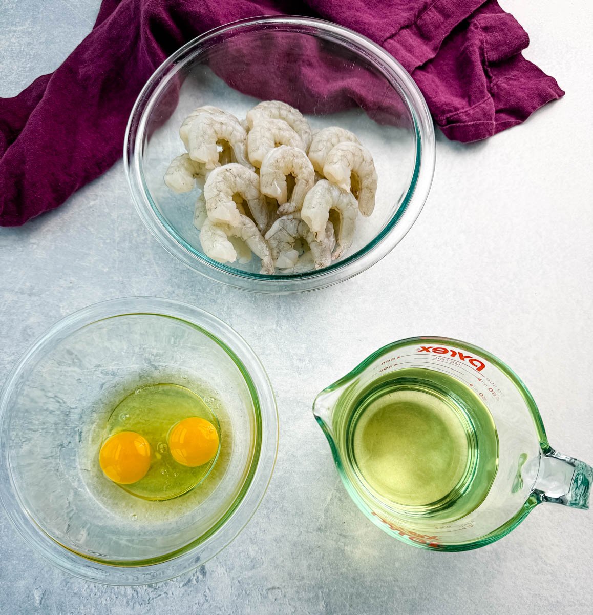 Overhead shot of prep ingredients for fried shrimp including raw peeled shrimp, cracked eggs in a bowl, and oil in a measuring cup.
