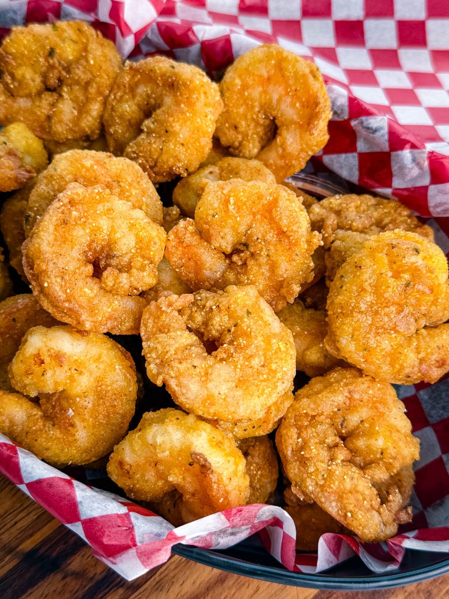 Fried shrimp in a basket lined with red and white paper, topped with hot sauce, with bowls of tartar sauce and cocktail sauce behind the basket.