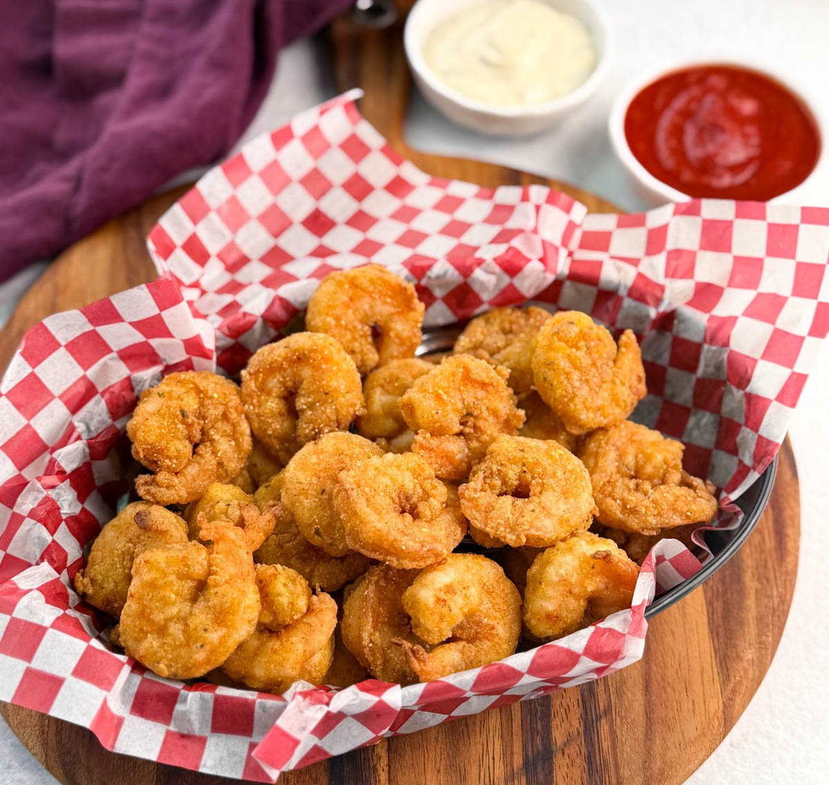 Front-focused shot of crispy fried shrimp piled high in a checkered basket, with sauce cups slightly out of focus in the background.