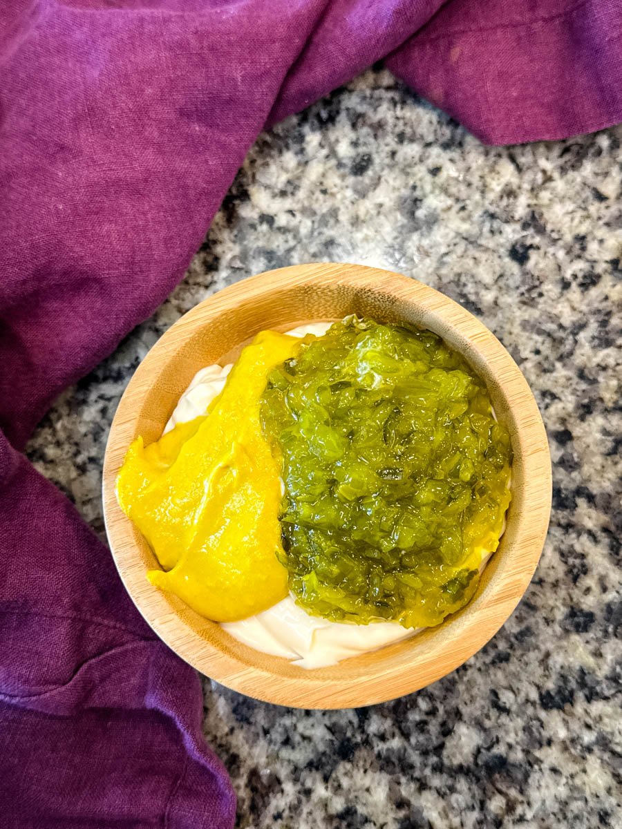 A wooden bowl showing unmixed layers of mayo, yellow mustard, and sweet pickle relish side by side, sitting on a granite counter.