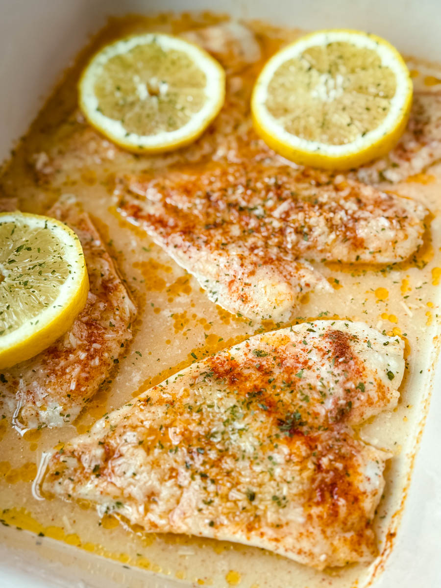 Close-up of golden baked garlic Parmesan flounder fillets with lemon slices and herbs in a buttery pan sauce.