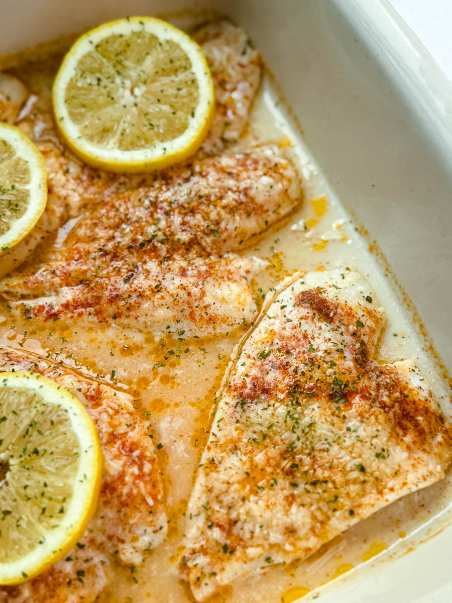 Cooked flounder fillets in a baking dish, lightly golden with melted Parmesan and seasoning, garnished with lemon slices and sprinkled with herbs.