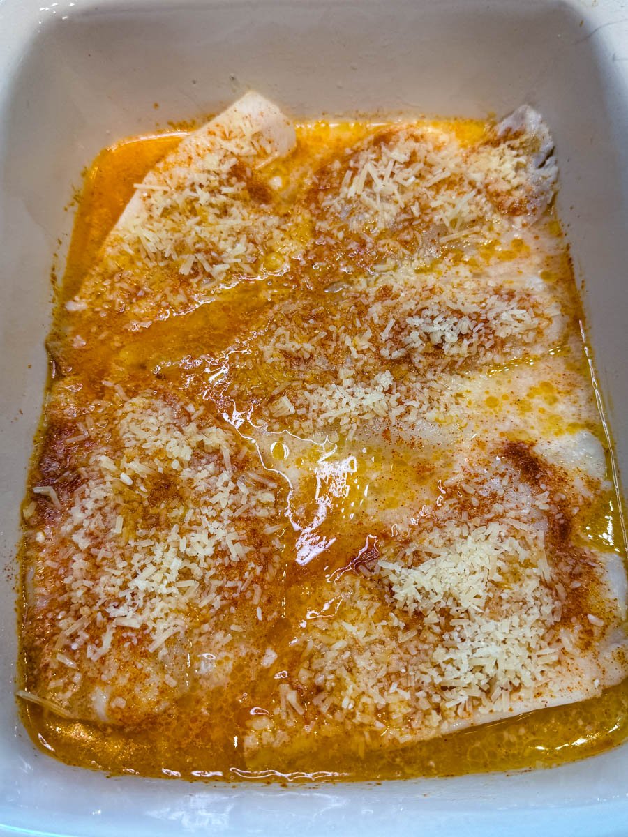 Unbaked flounder fillets in a dish with butter, seasonings, and a topping of shredded Parmesan cheese just before going into the oven.