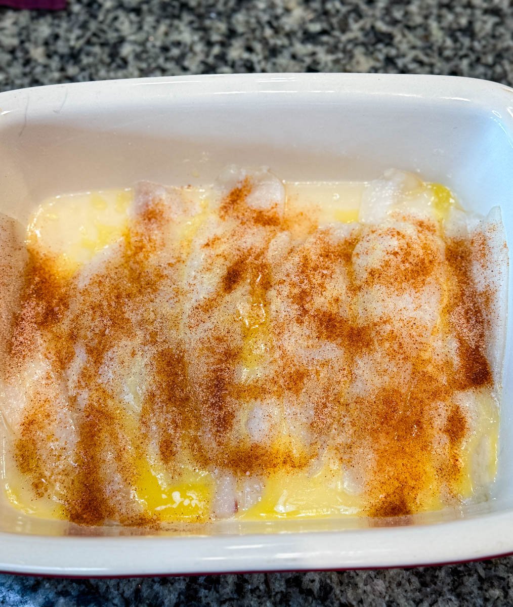 Flounder fillets in a white baking dish sprinkled with a generous layer of seasoning including paprika and garlic powder over melted butter.