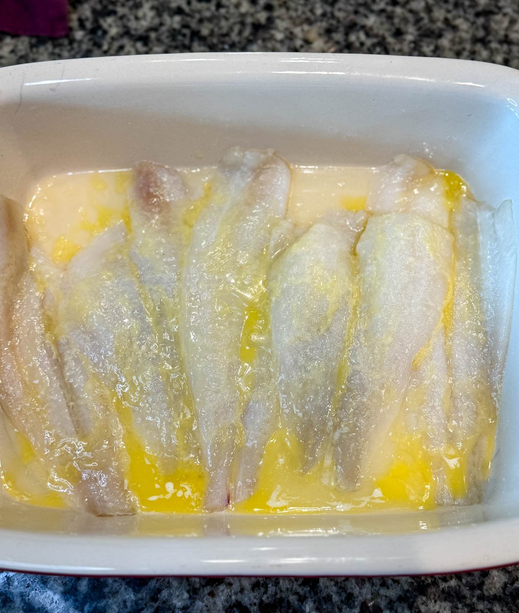 Flounder fillets placed in a white baking dish, coated with melted butter before seasoning and baking.