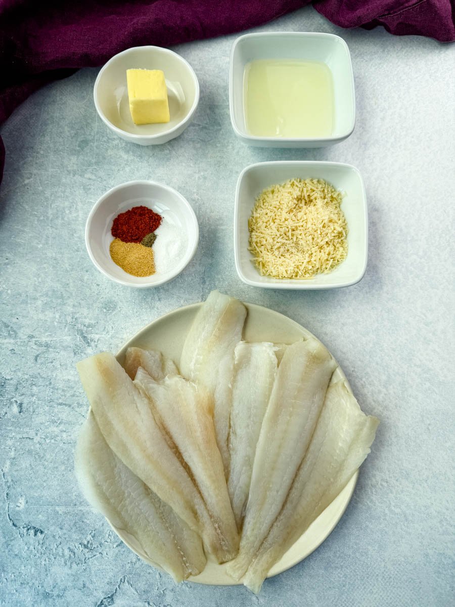 Overhead view of ingredients for garlic Parmesan flounder, including raw flounder fillets, grated Parmesan cheese, butter, olive oil, and a spice mix of paprika, garlic powder, salt, and pepper, all arranged on a light surface.