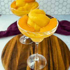 Two cocktail glasses filled with thick peach bellini slush, garnished with peach slices, placed on a wooden board.