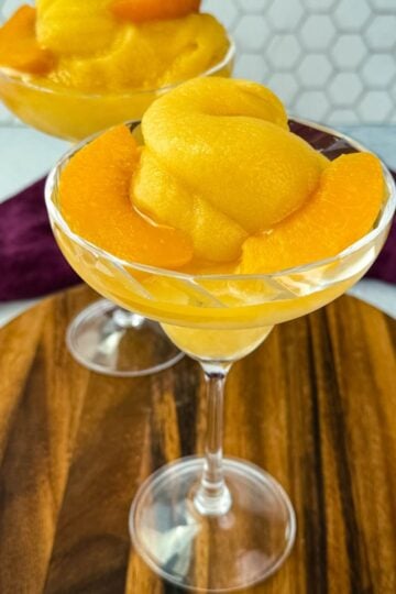 Two cocktail glasses filled with thick peach bellini slush, garnished with peach slices, placed on a wooden board.