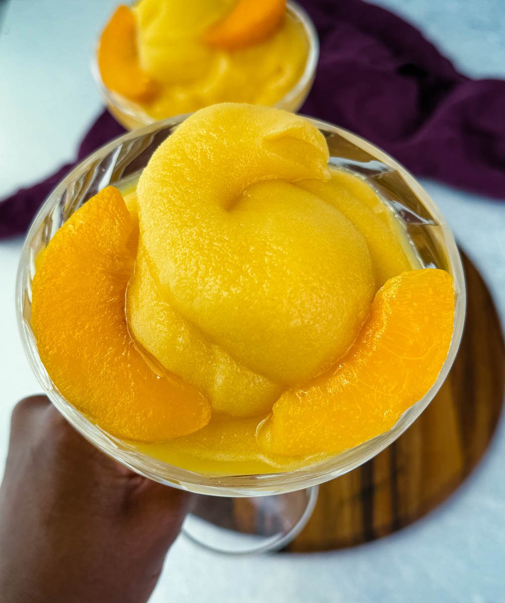 Close-up of a hand holding a glass filled with creamy frozen peach bellini slush and peach slice garnish.