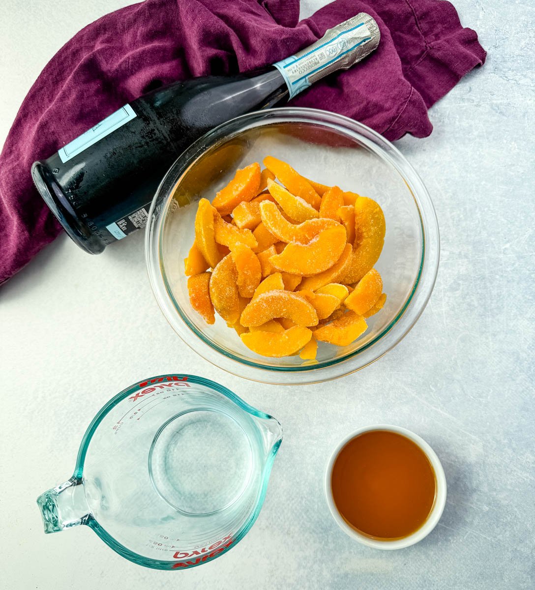 Ingredients for a peach bellini slush laid out on a countertop: frozen peach slices in a bowl, a bottle of Prosecco, peach nectar, and water.