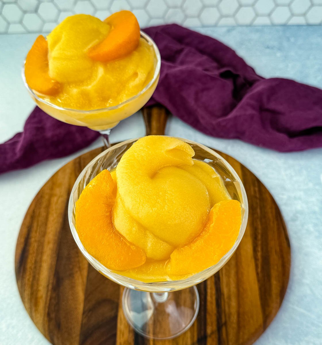Two cocktail glasses filled with thick peach bellini slush, garnished with peach slices, placed on a wooden board.