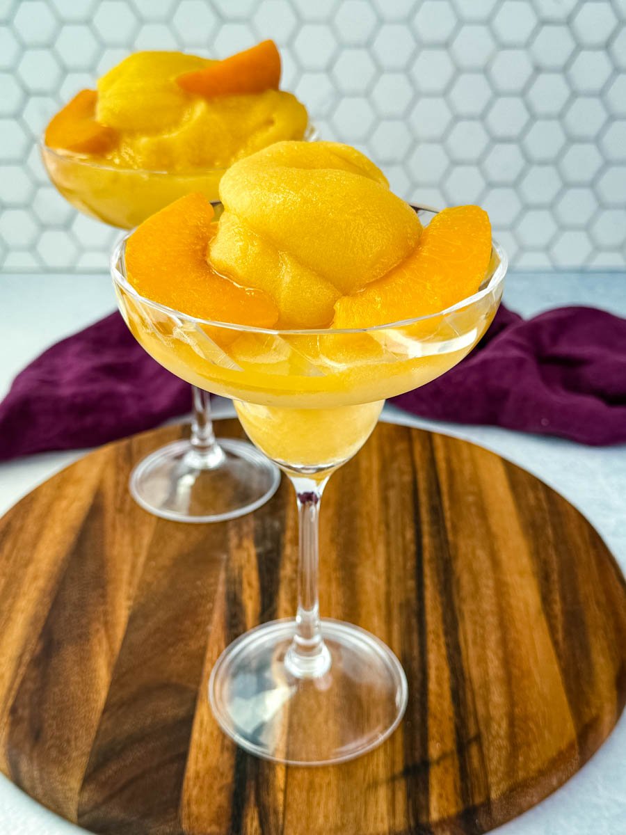 Two cocktail glasses filled with thick peach bellini slush, garnished with peach slices, placed on a wooden board.