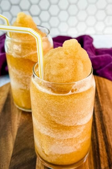 Closer side angle of iced tea slush in two glasses with yellow-striped straws on a wooden board.