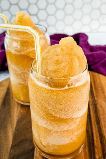 Closer side angle of iced tea slush in two glasses with yellow-striped straws on a wooden board.