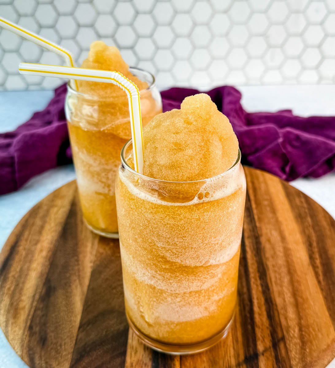 Side angle of the iced tea slush in two clear glasses with straws, against a white tile background.