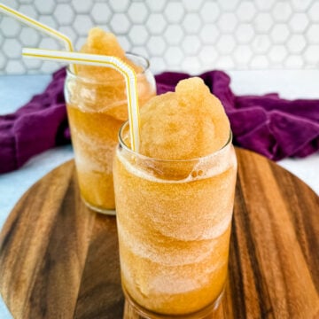 Closer side angle of iced tea slush in two glasses with yellow-striped straws on a wooden board.