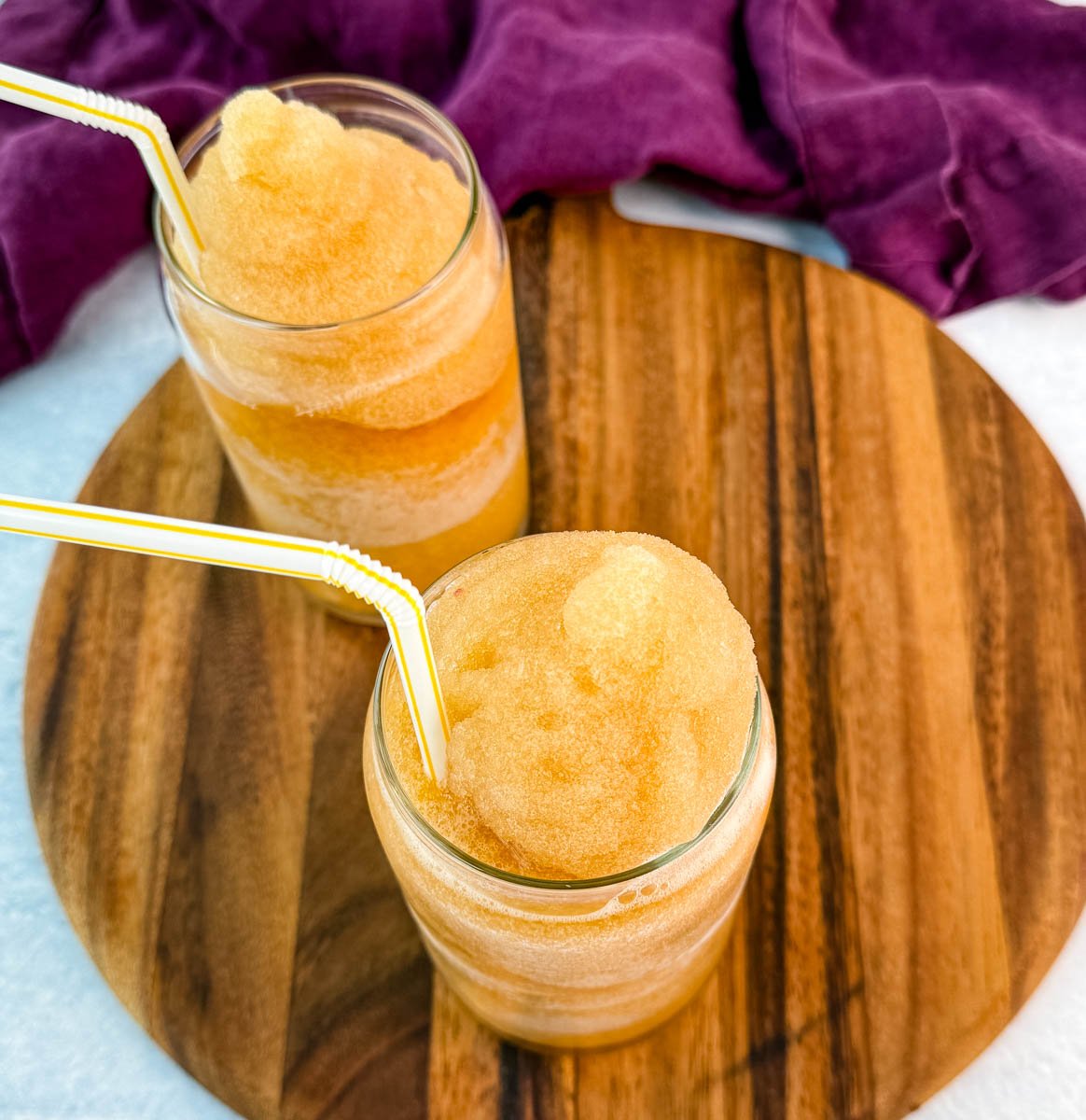 Two glasses of iced tea slush on a round wooden board, each with a yellow-striped straw.