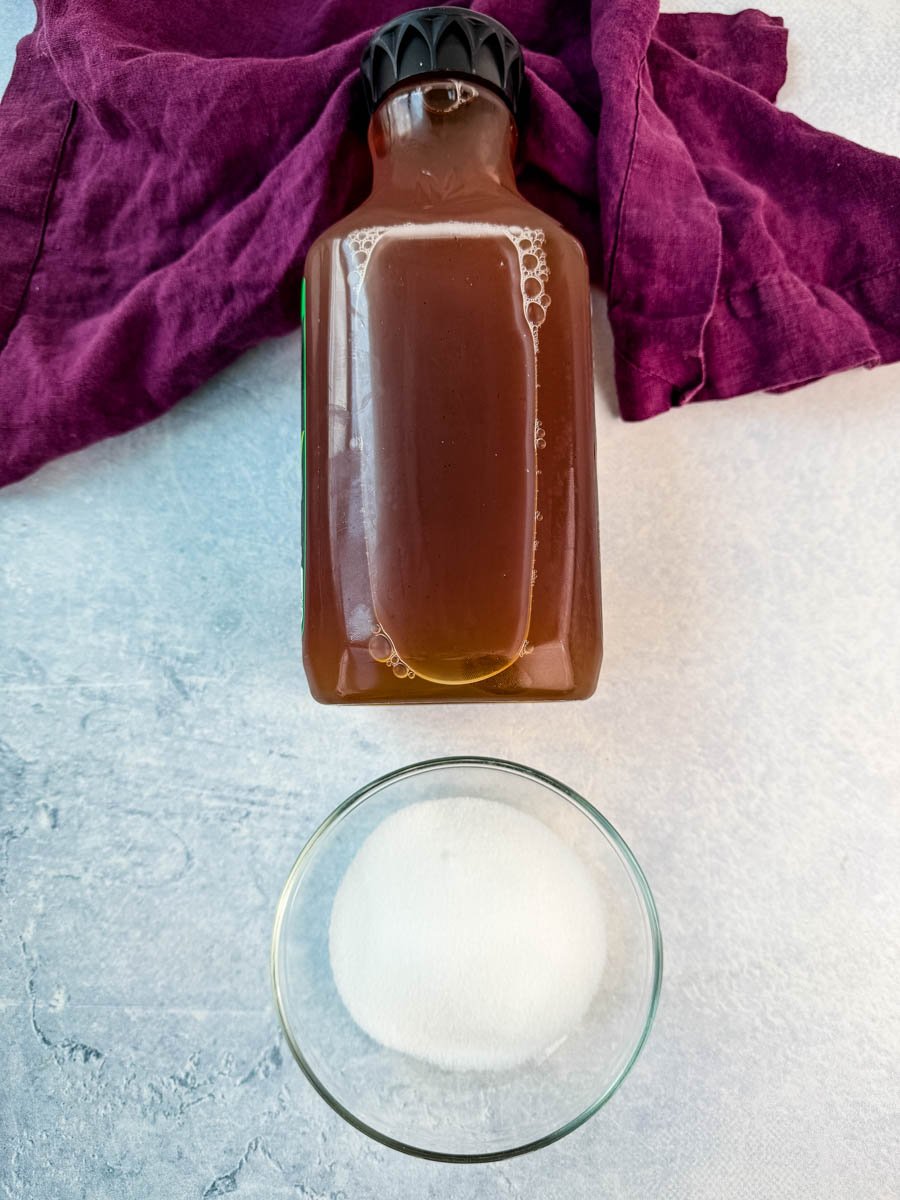 A bottle of iced tea and a small glass bowl of white sugar on a white surface with a purple cloth napkin.