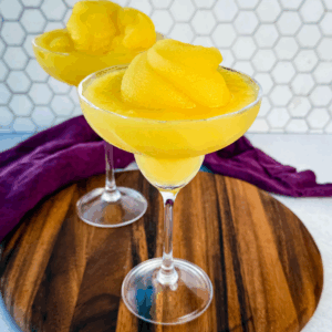 Two frozen mimosas in coupe glasses sitting on a wooden board with a hexagon tile backsplash in the background.