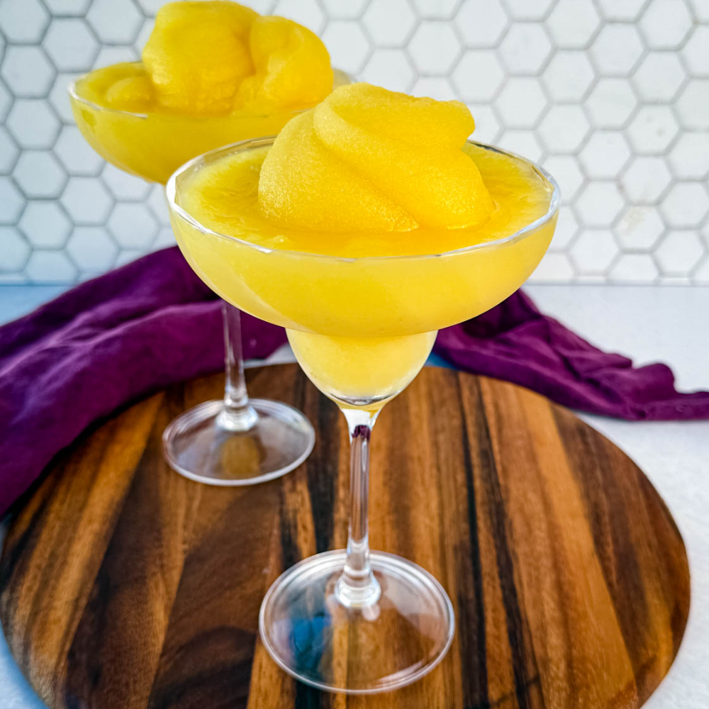 Two frozen mimosas in coupe glasses sitting on a wooden board with a hexagon tile backsplash in the background.