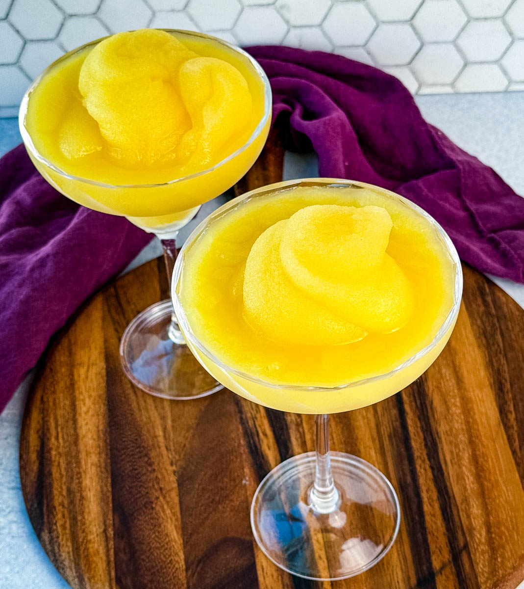 Angled view of two frozen mimosa cocktails garnished with their signature slush swirl, styled on a wooden surface with a purple cloth.