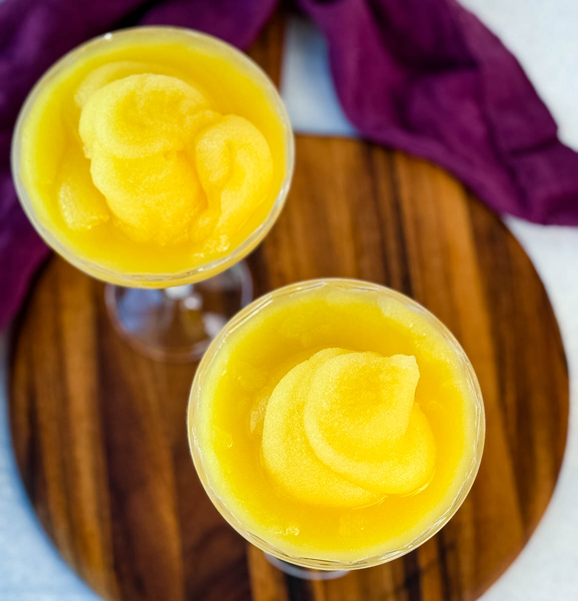 Overhead shot of two coupe glasses filled with swirled frozen mimosa slush on a wooden board.