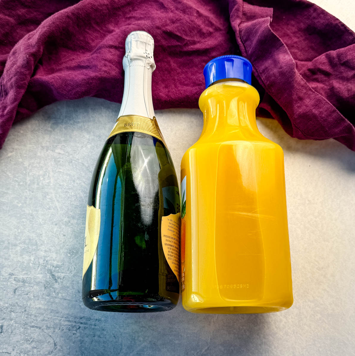 A bottle of champagne and a plastic bottle of orange juice on a light surface with a dark purple linen in the background.