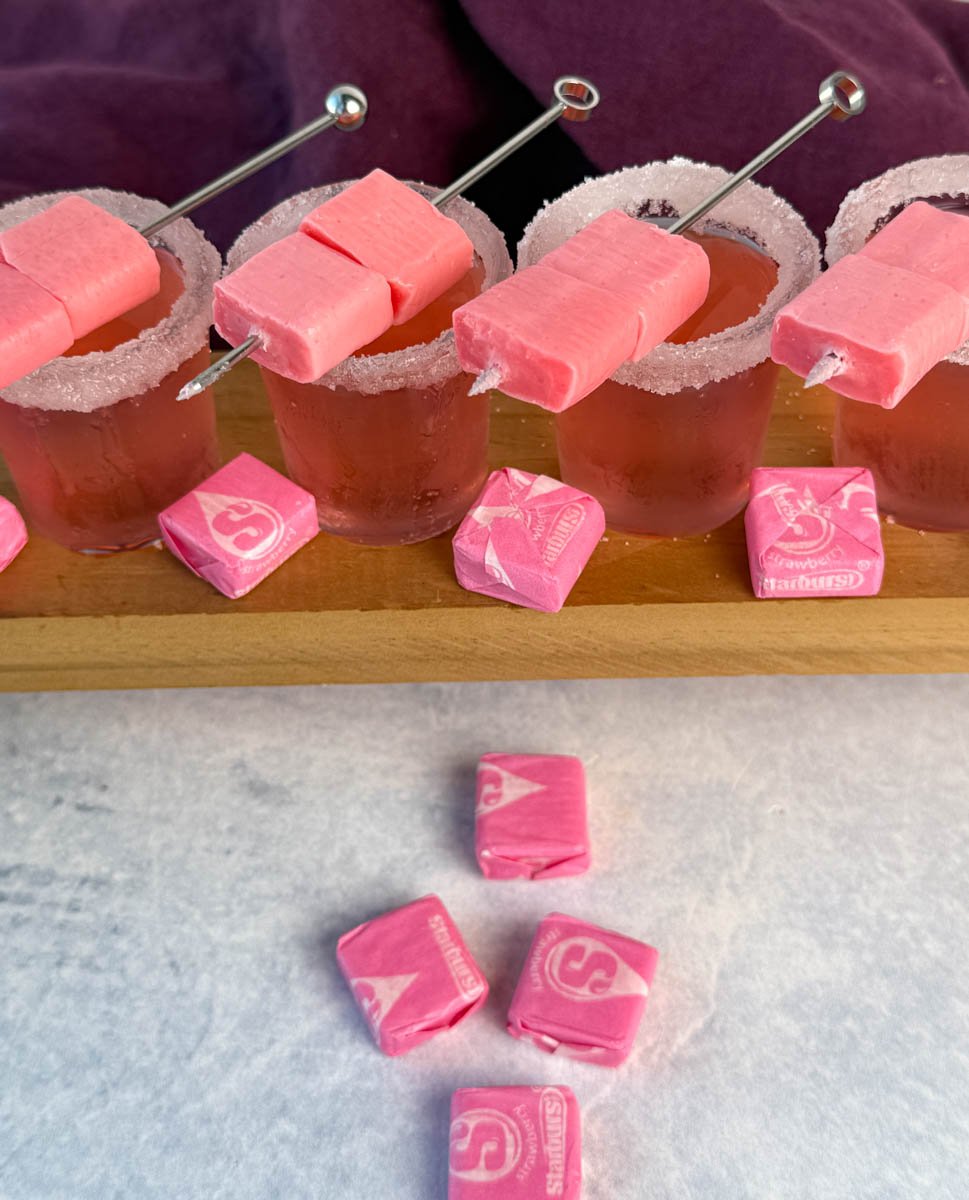 Overhead view of four pink Starburst shots with sugared rims and skewered pink Starbursts, surrounded by wrapped candy.