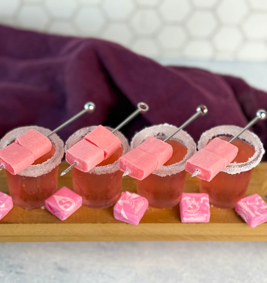 Overhead view of four pink Starburst shots with sugared rims and skewered pink Starbursts, surrounded by wrapped candy.