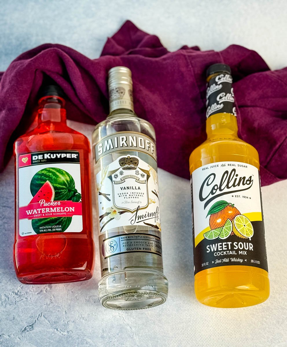 Bottles of DeKuyper Watermelon Schnapps, Smirnoff Vanilla Vodka, and Collins Sweet & Sour Mix arranged next to a purple kitchen towel.