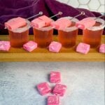 Four pink Starburst-inspired cocktail shots with sugar-rimmed glasses, each garnished with skewered pink Starburst candies. More pink Starbursts are scattered on the table in front of the drinks, with a hexagon-tiled wall and deep purple cloth in the background.