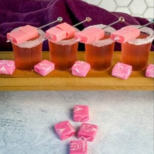 Four pink Starburst-inspired cocktail shots with sugar-rimmed glasses, each garnished with skewered pink Starburst candies. More pink Starbursts are scattered on the table in front of the drinks, with a hexagon-tiled wall and deep purple cloth in the background.