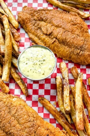 Golden fried catfish served with hand-cut fries and a side of creamy tartar sauce, all arranged on red and white checkered paper in a black tray.
