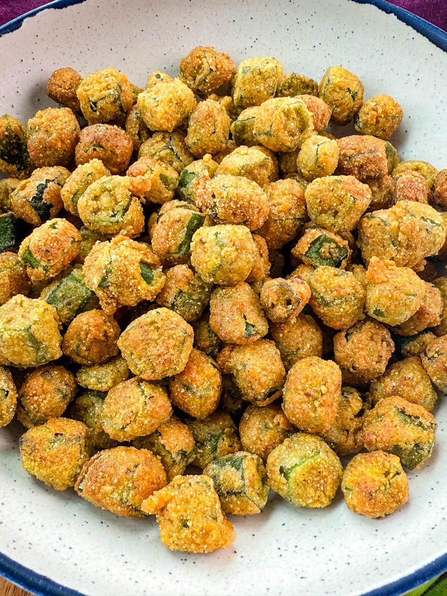 Crispy fried okra served in a large white and blue speckled bowl on a wooden surface with a purple cloth in the background.