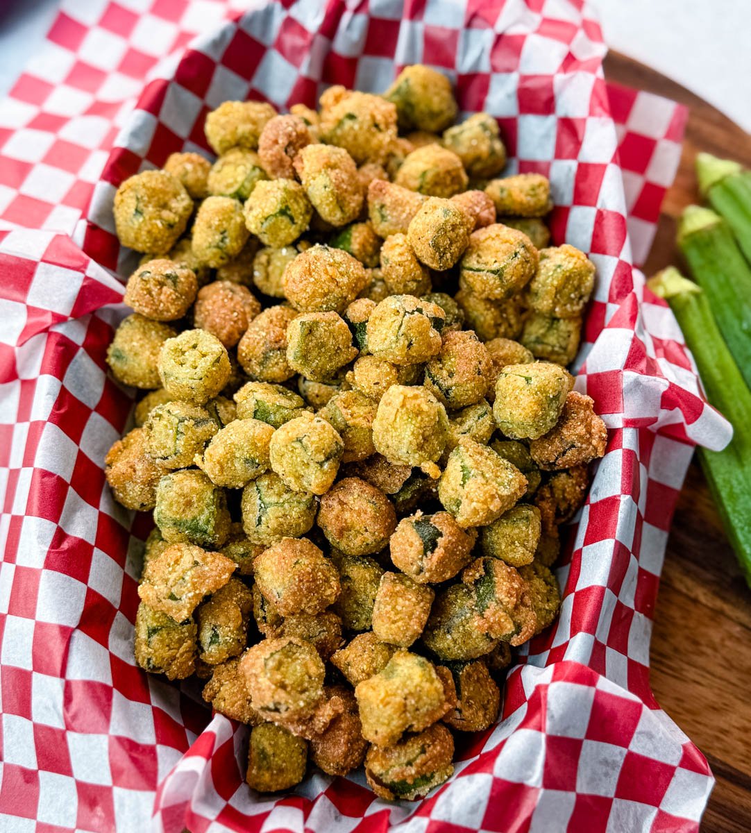 Crispy Southern fried okra in a red and white checkered basket, styled on a wood board with fresh okra pods next to it
