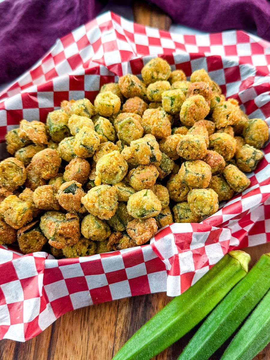 Front view of a basket filled with perfectly crisp fried okra on red-checkered paper, with fresh okra pods resting nearby.