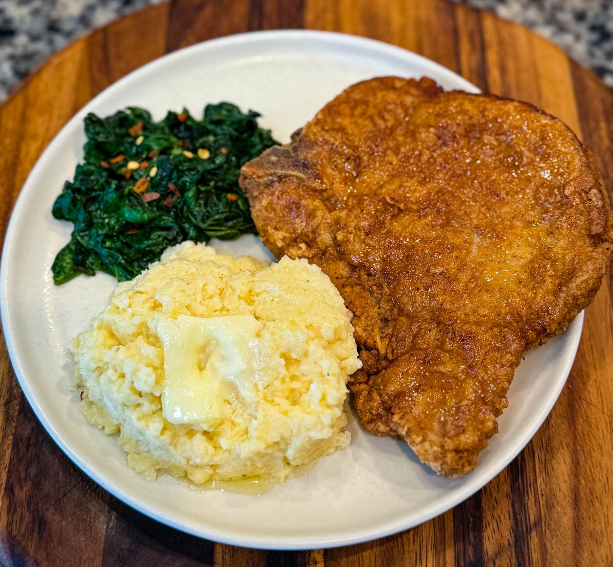 A plate of fried pork chop served with buttery cheese grits and sautéed greens.