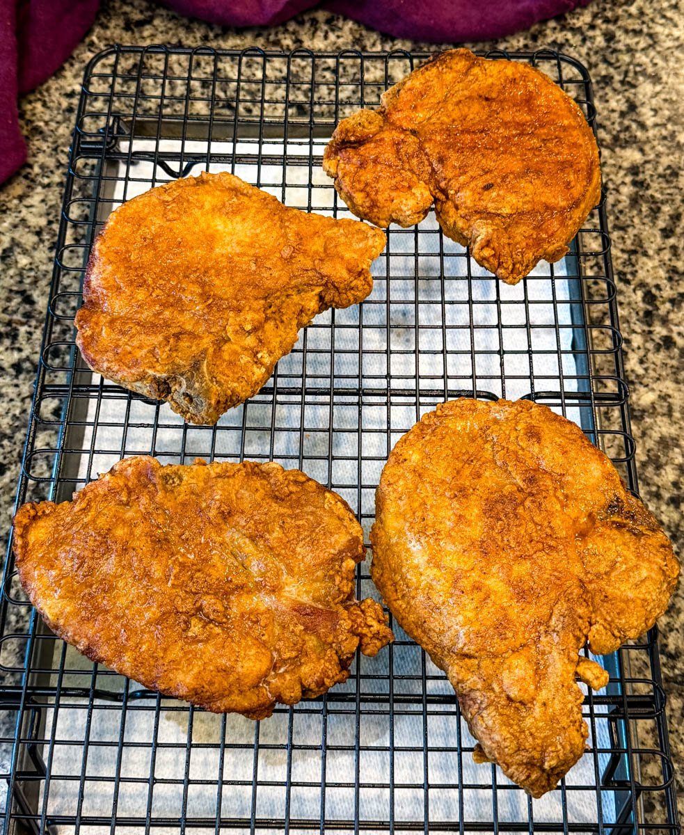 Four freshly fried pork chops resting on a wire rack to cool and stay crispy.
