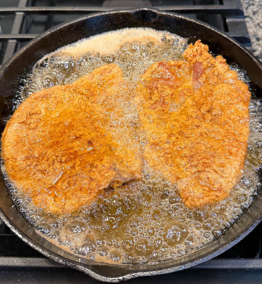 Two bone-in pork chops frying in hot oil in a cast iron skillet, with visible bubbles around the edges.