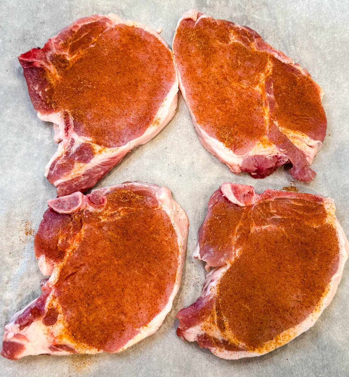 Four raw bone-in pork chops seasoned with spices, arranged on parchment paper.