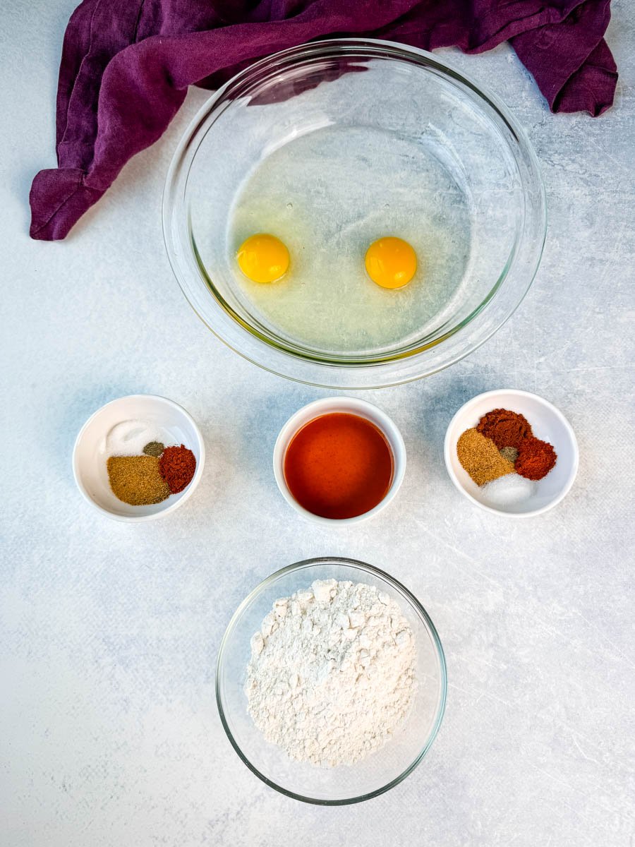 Overhead shot of ingredients for fried pork chops, including two eggs in a bowl, flour, hot sauce, and various spices in small bowls.