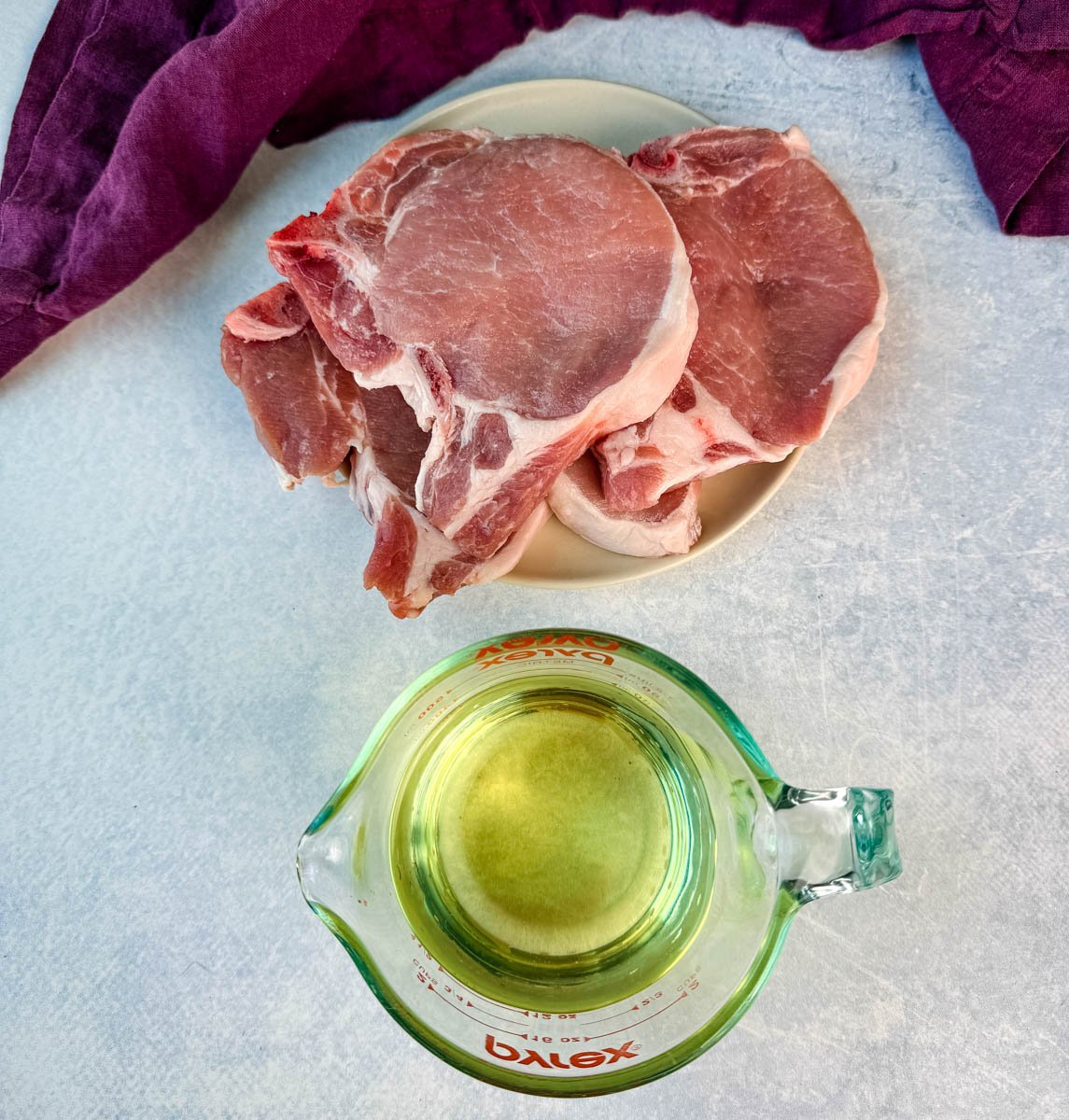 A plate of raw bone-in pork chops next to a measuring cup filled with oil, prepped for frying.