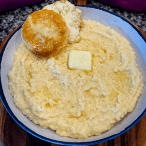 Close-up of creamy stone ground cheese grits in a speckled bowl, topped with a pat of melting butter and a flaky biscuit on the side.