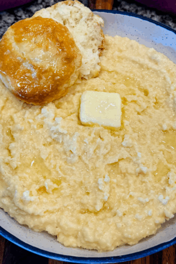 Close-up of creamy stone ground cheese grits in a speckled bowl, topped with a pat of melting butter and a flaky biscuit on the side.